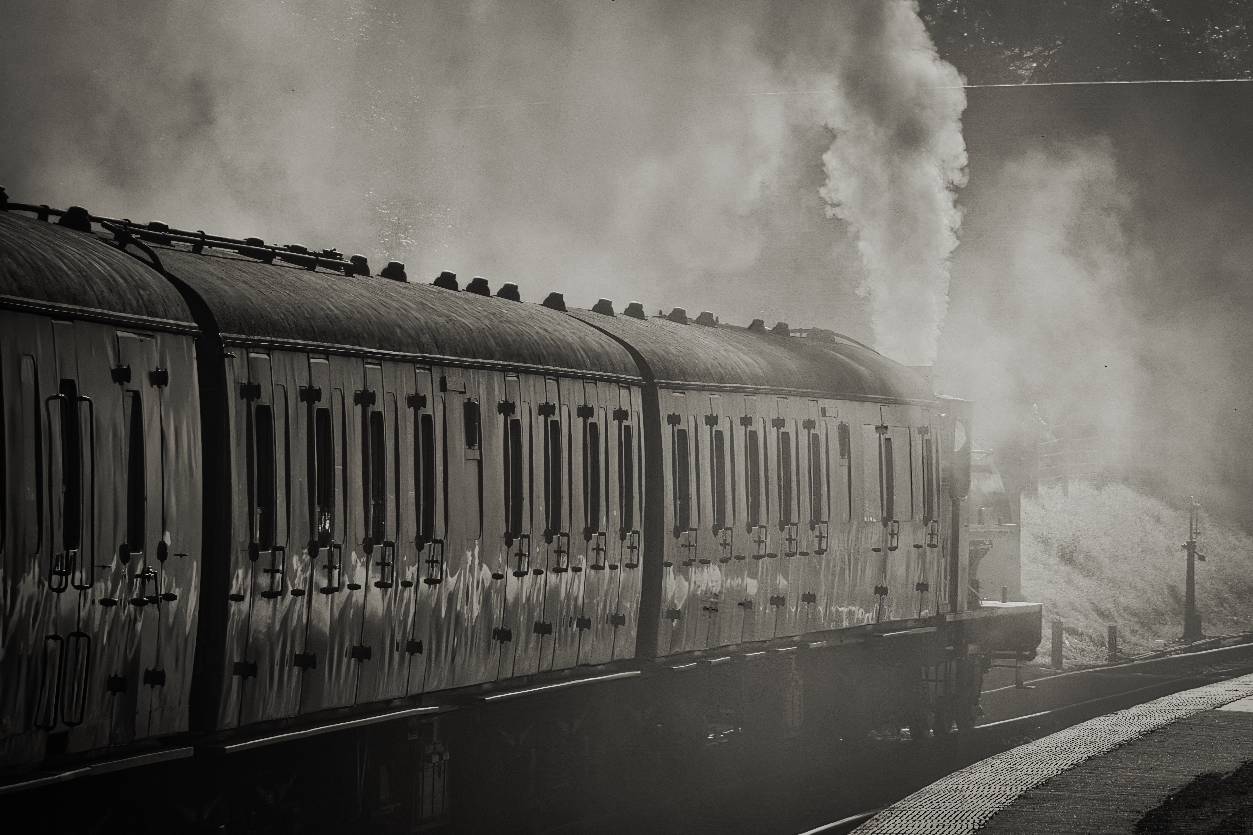 This evocative photograph captures the essence of a steam train in moment it's in midst of chugging along, with billowing smoke adding a dramatic flair. The monochromatic palette enhances the vintage charm, while the composition draws the eye along the length of the train, creating a perfect balance of movement and stillness. The interplay of light and shadow, combined with the textured details of the train's surface, makes this image a perfect piece for a modern gallery wall or a executive office, where it can serve as a focal point, evoking a spirit of adventure and nostalgia.