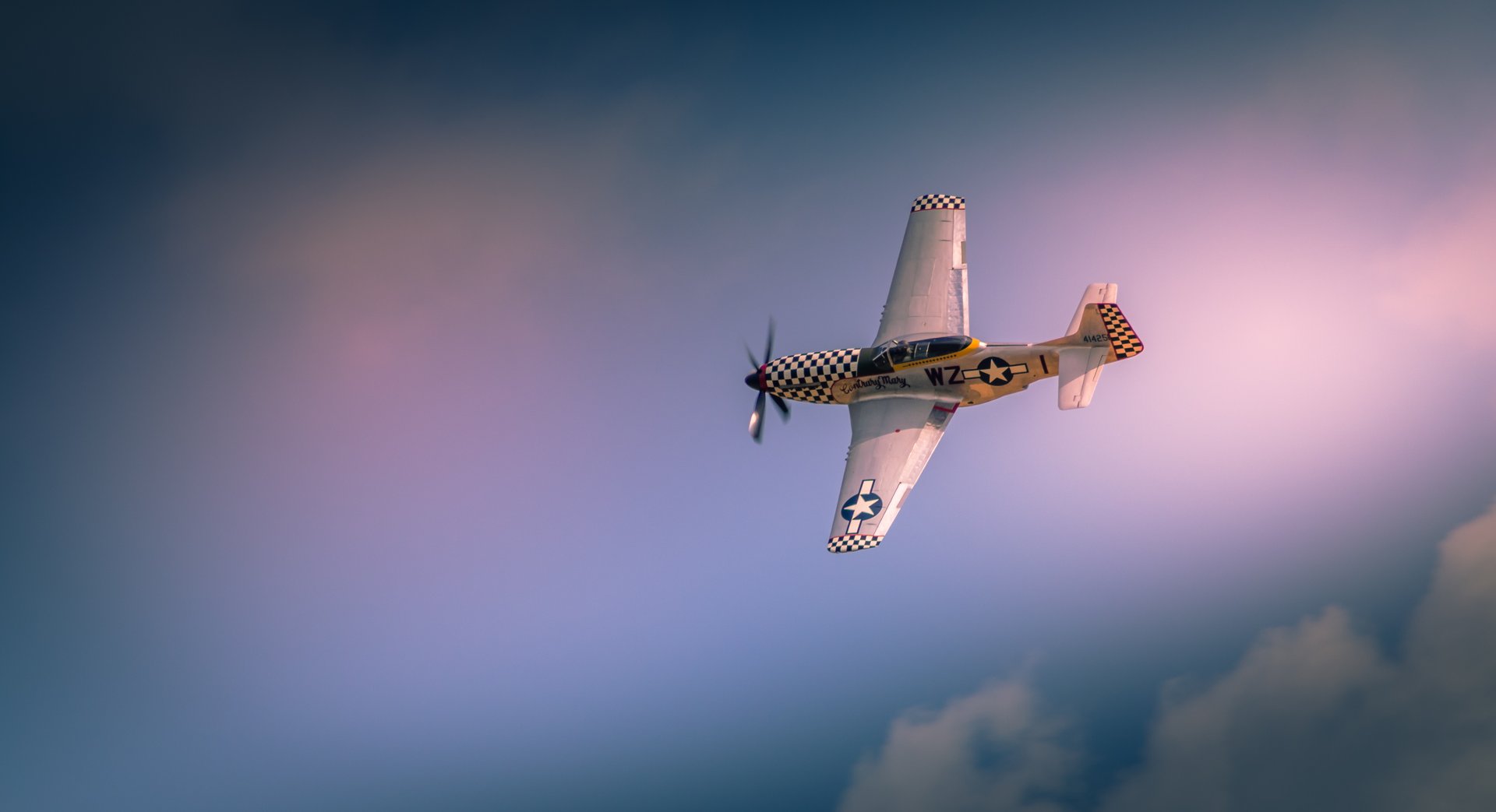 Capture the essence of classic aviation with this stunning photograph of a P-51 Mustang in flight. The image, bathed in the warm glow of a setting sun, evokes a spirit of adventure and freedom. The checkered tail and star emblem add a perfect touch of vintage charm, making it a captivating piece for any aviation enthusiast. Perfect for a living room as a a focal point, or as a a centerpiece of a modern gallery wall in an executive office, this photograph is a timeless addition to any decor.